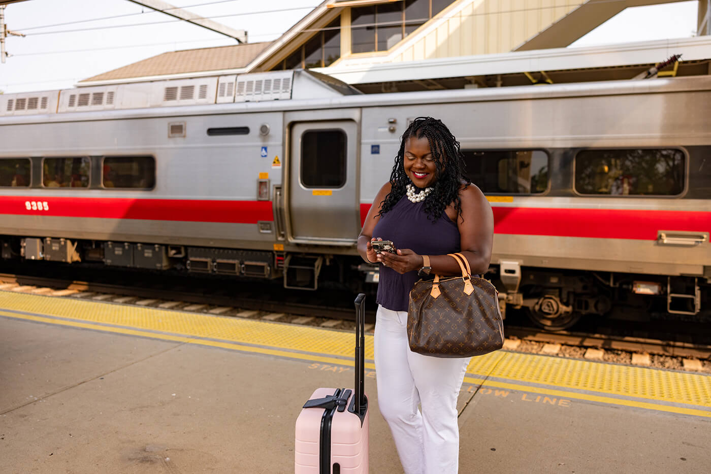 A woman stands in front of a train inside a city station. She is looking at her phone while waiting on the platform with her luggage.